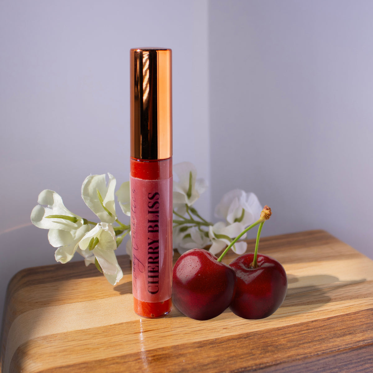 A tube of Liney Cosmetics Cherry Bliss gloss with cherries sitting next to it and white flowers in the background.