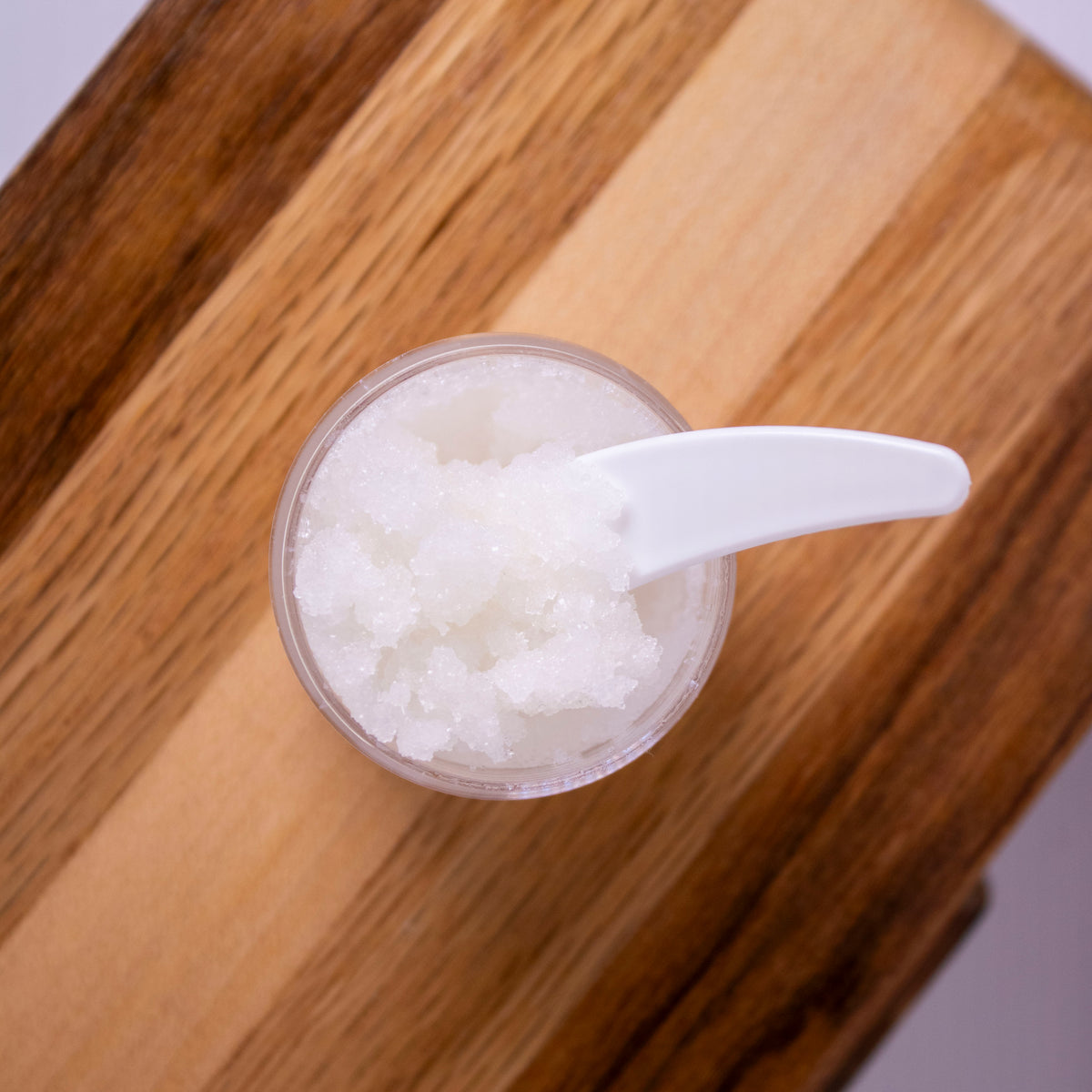 A small jar of Liney Cosmetics Lip Scrub, with a white plastic spoon resting inside, placed on a wooden board.