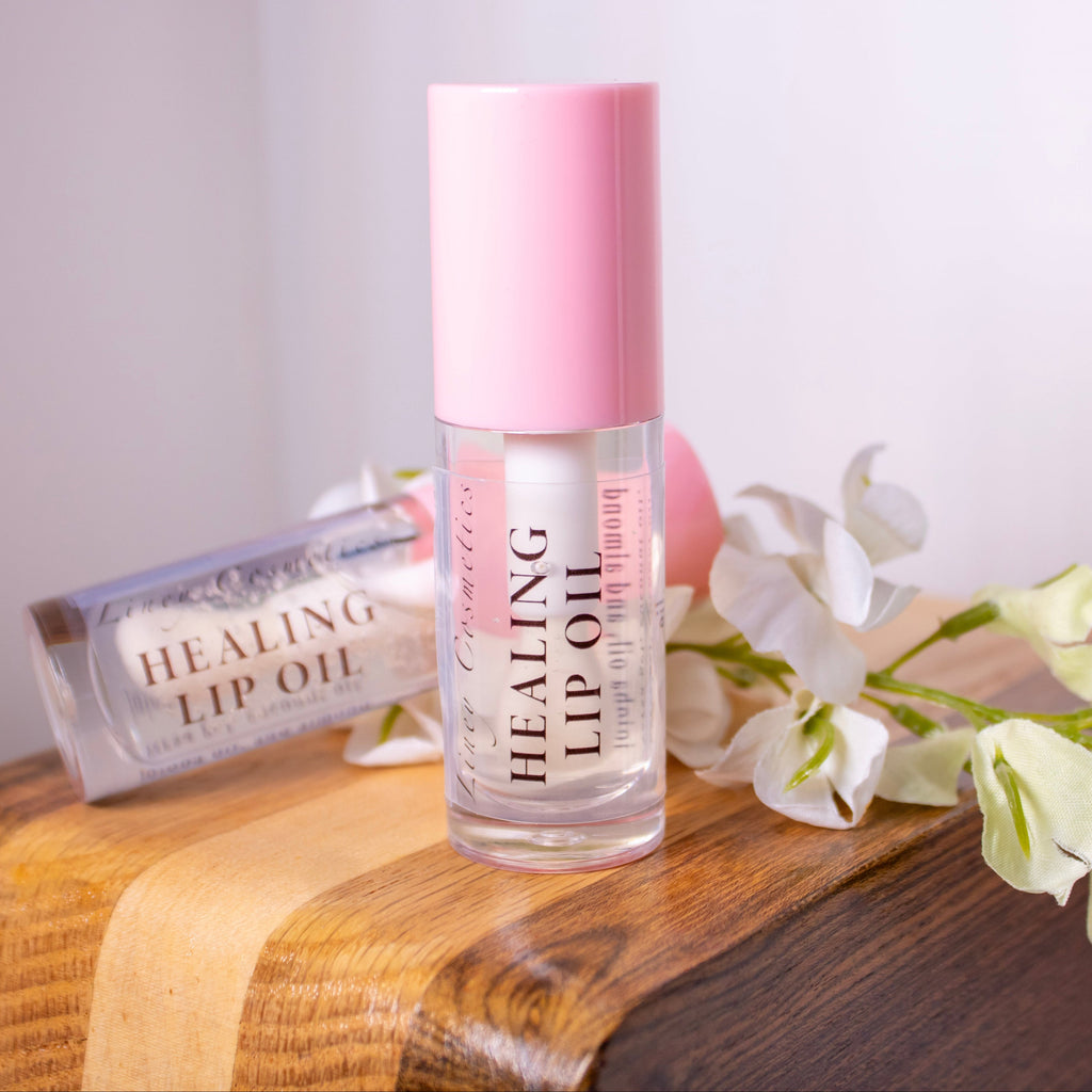 A bottle of Liney Cosmetics Healing Lip Oil with a pink cap stands on a wooden surface next to a matching lip oil tube, with white flowers beside it.