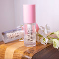 A bottle of Liney Cosmetics Healing Lip Oil with a pink cap stands on a wooden surface next to a matching lip oil tube, with white flowers beside it.