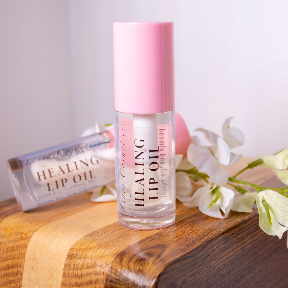 A bottle of Liney Cosmetics Healing Lip Oil with a pink cap stands on a wooden surface next to a matching lip oil tube, with white flowers beside it.