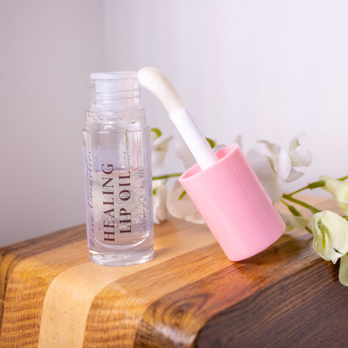 A bottle of Liney Cosmetics Healing Lip Oil with its applicator resting on the side on a wooden surface next to a matching lip oil tube, with white flowers beside it.