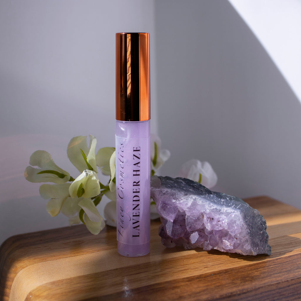 A tube of Liney Cosmetics Lavender Haze Gloss sitting on wood with a purple crystal next to it and white flowers in the background.
