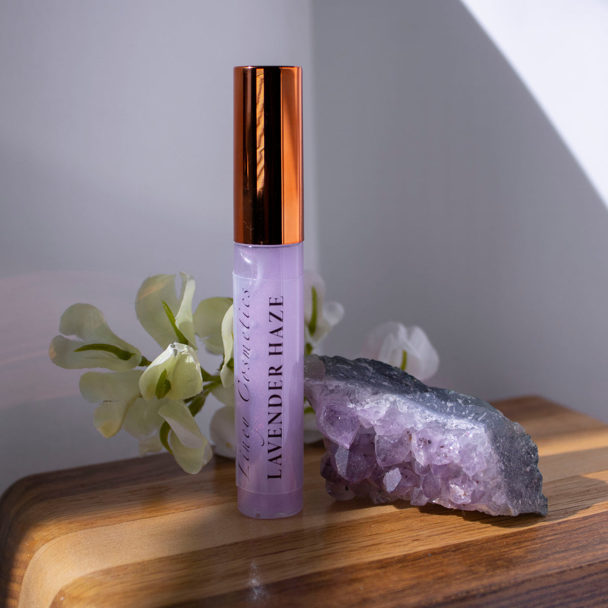 A tube of Liney Cosmetics Lavender Haze Gloss sitting on wood with a purple crystal next to it and white flowers in the background.