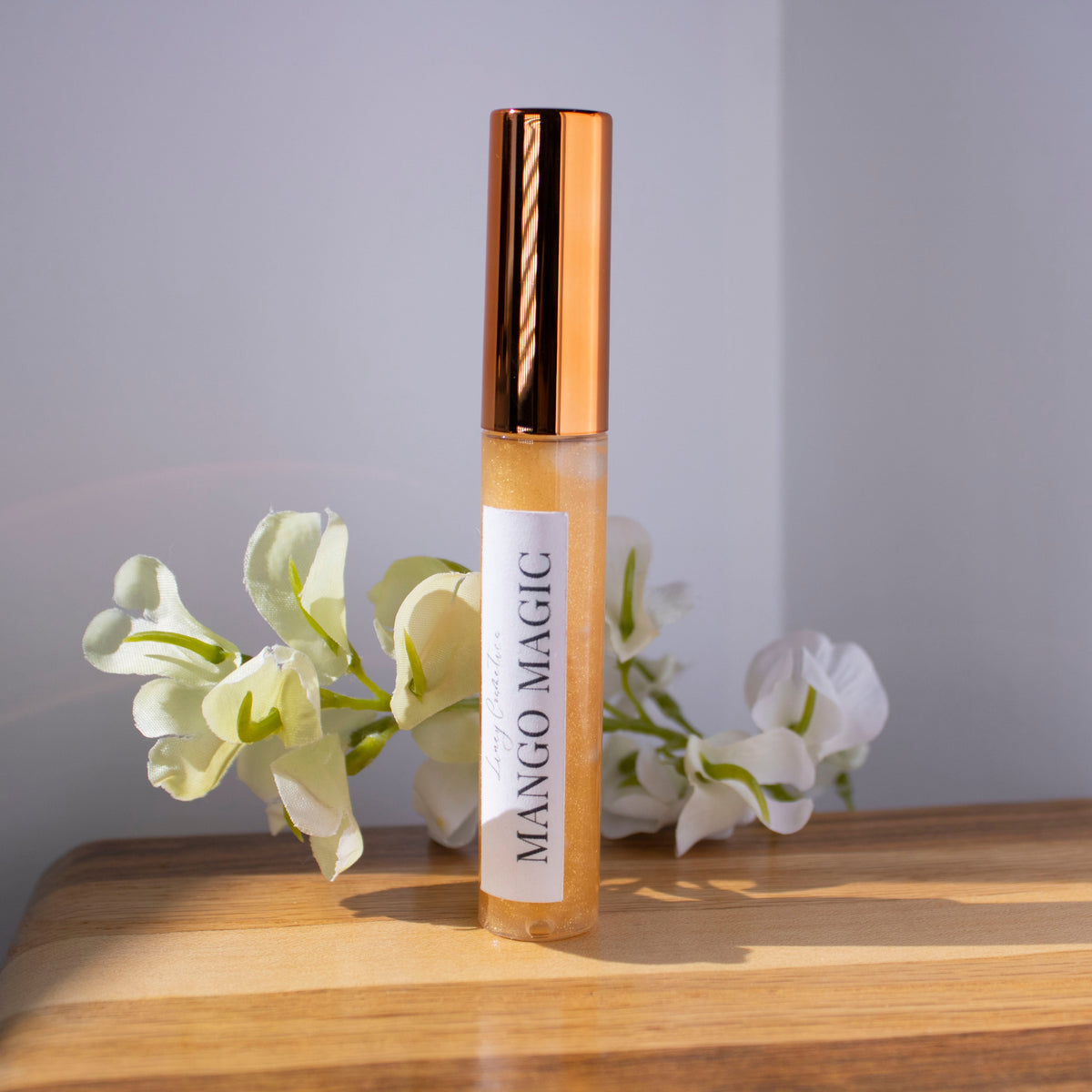 A tube of Liney Cosmetics Mango Magic Gloss sitting on a wood with white flowers in the background.
