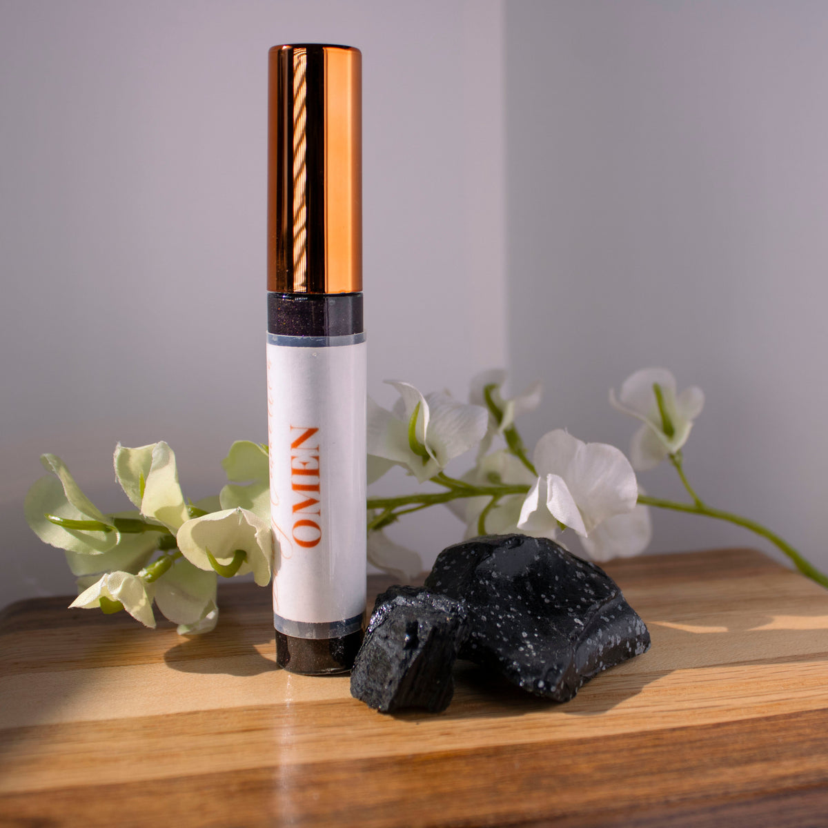 A tube of Liney Cosmetics Omen Gloss sitting on wood with obsidian next to it and white flowers in the background.