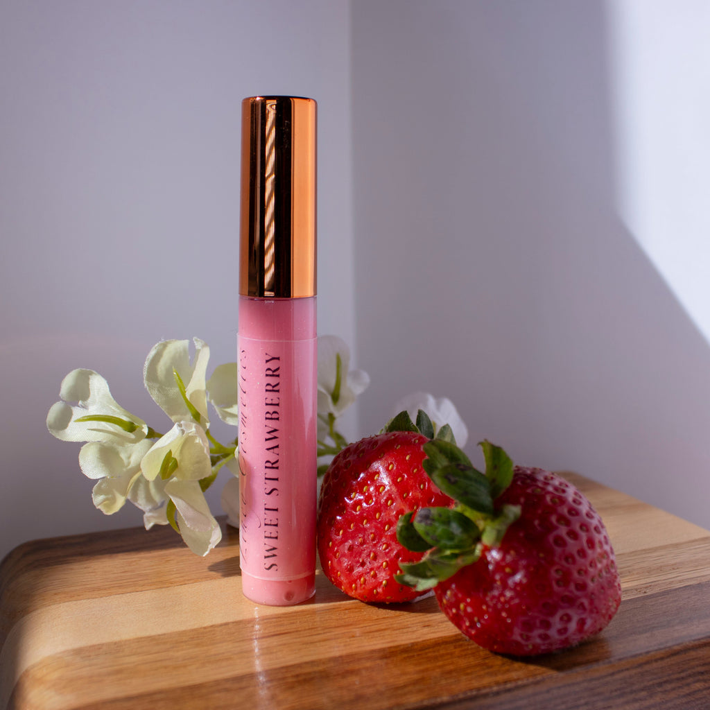 Liney Cosmetics Sweet Strawberry gloss with strawberries sitting next to it on a wooden surface, with white flowers in the background.