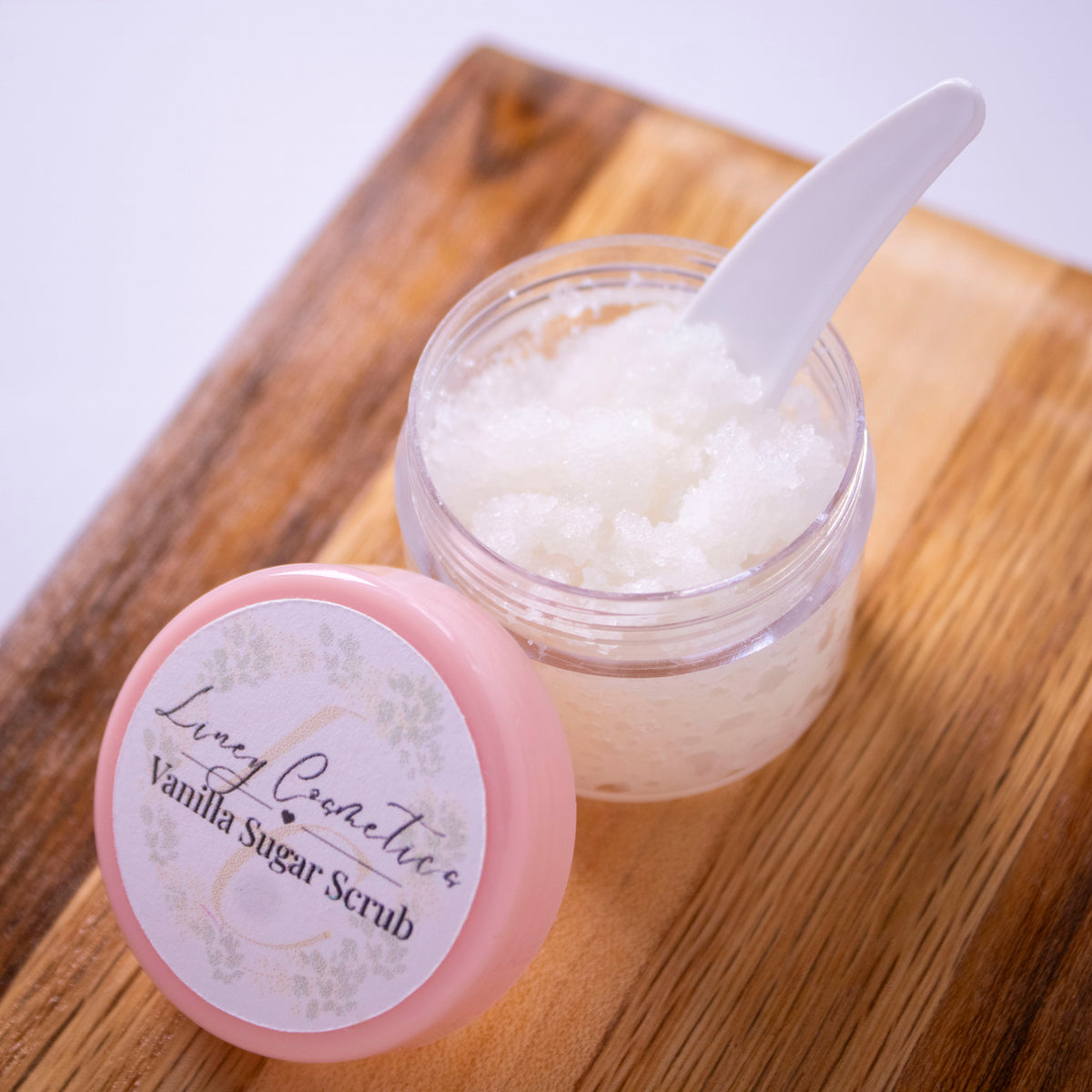 A jar of Liney Cosmetics Vanilla Lip Scrub with its pink lid resting beside it on a wooden cutting board.